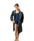 Child standing wearing a black swim parka. Black swim parka has cotton towel lined, showerproof outer and pockets. Child has her hand in the pocket and swimmers on.