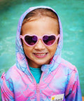 Child wearing pink love heart sunglasses standing in front of a pool wearing a schmik swim parka in mermaid print. 