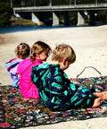 3 children sitting on a wandering folk rug wearing schmik swim parkas. Scene is on the beach in currumbin and one child wears a mermaid print, fluro pink and jellyfish  print swim parka. 
