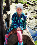 Child sitting on a log wearing a schmik swim parka. Child is camping and wears the jacket to keep warm.