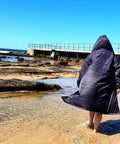 Adult wearing a black Schmik swim parka. Adult is walking on the beach and has the swim parka on with he hood on thier head. Adult is walking on the beach.