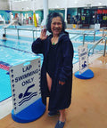 Happy asain lady wearing a black swim parka. Lady is standing in front of a lap swimming only sign. Black swim parka is long and made to be used after swimming.