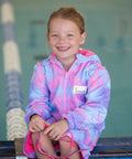 Child sitting next to pool wearing mermaid print swim parka. Mermaid print swim parka is pink and comes with removable sleeves. 