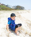 Boy sitting on the beach on the Gold Coast weaing a swim parka. Swim parka has its arms removed and is fishbone print.