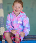 Child sitting on edge of pool. Child is wearing mermaid print swim parka and is holding goggles. Mermaid print swim parka is pink with mermaid sclales printed all over it. 