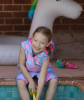 Child sitting on edge of swimming pool wearing pink mermaid print swim parka. 