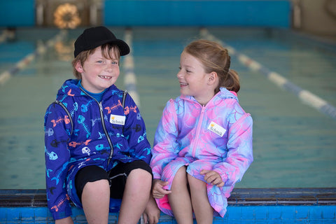 Boy and girl sitting on pool side wearing blue fishbone print swim parka and pink mermiad print swim parka. Schmik swim jackets. 