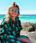 Child sitting on rock at the beach wearing a jellyfish print swim parka. Schmik jellyfish print swim parka is navy and green with green jellyfish printed all over it. 